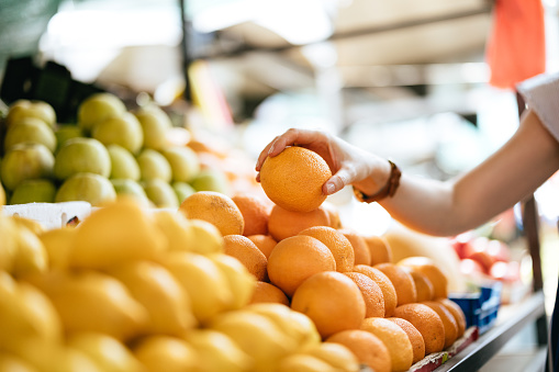 Zoomed in photo of an unrecognizable woman picking up and buying fruit. She is holding an orange.