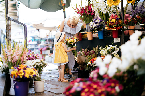 Beautiful woman in a dress standing in the flower shop with her young daughter who is turned away from the camera. They are picking flowers to buy. Mother is looking down at the daughter and smiling while the girl is holding a bouquet of flowers.