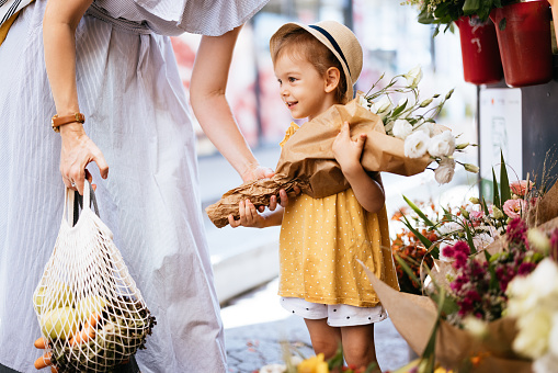 Portrait of a cute little girl in a yellow shirt and a hat holding a bouquet of white flowers. She is looking away and smiling. Her anonymous mother is nearby.