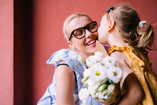 Beautiful happy woman is crouching while her unrecognizable young girl is holding a bouquet of white flowers and kissing her on the cheek. She is smiling with her eyes closed.