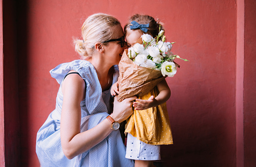 Beautiful happy woman is holding a bouquet of white flowers while hugging her unrecognizable young girl. She is smiling while looking at her daughter.