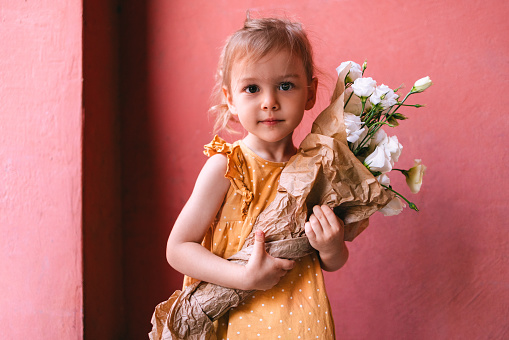Close up shot of a cute young girl in a yellow dress holding a bouquet of white flowers. There is a red wall behind her.