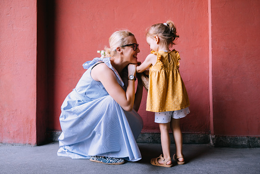 Beautiful cheerful woman in a blue dress crouching and looking at her young daughter standing in front of her. She is smiling and holding her little arms. The anonymous girl is looking down at her mom.