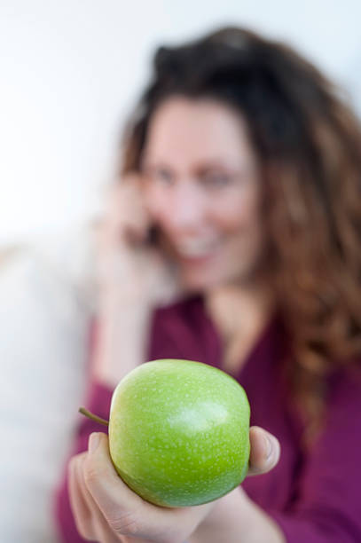 Vertical shot of a happy smiling young woman in violet shirt offering a green apple while talking on her mobile phone. Shallow DOF, focus on the apple in the foreground