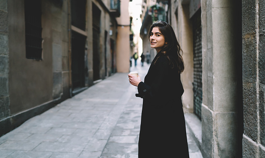 Half length portrait of attractive female tourist with takeaway cup looking at camera during coffee time on sightseeing, beautiful Caucasian girl 20 years old posing while exploring city streets