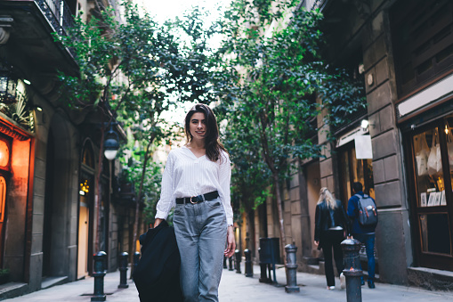 Half length portrait of attractive female tourist dressed in casual clothing enjoying travelling vacations, charming hipster girl looking at camera while walking at town street during getaway journey