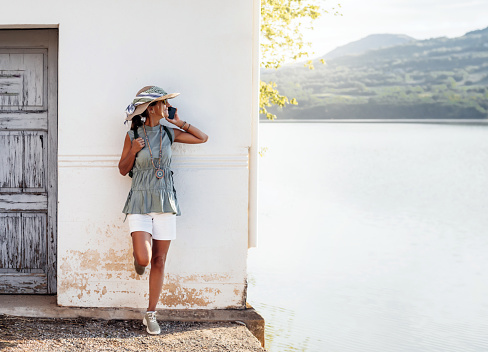 a woman wearing a hat and scarf, leans on the facade of a lakeside house and talks on the phone, while smiling. concept of fun and nature.