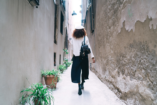 Back view of woman in beret and in trendy clothes with backpack while walking along narrow city street among buildings