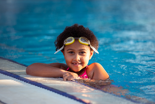 Portrait of multiracial elementary-age girl with curly hair wearing swim goggles swimming in a pool on a sunny day.