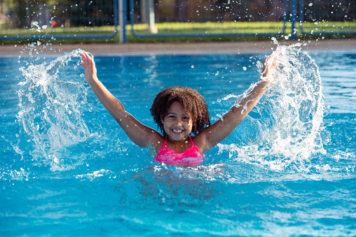 Happy curly hair little girl with wide open hands splashing water in the swimming pool. The girl having fun on summer vacation.