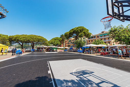 Lerici, Italy - July 16, 2022: Outdoor basketball court and street market in Lerici downtown. Tourist resort on the coast of the Gulf of La Spezia, Mediterranean Sea, Liguria, Italy, Europe. A large number of people stroll through the city market on a sunny summer day.