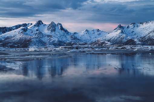 Frozen surface of sea reflecting rocky mountains with sharp peaks and snowy slopes against picturesque colorful cloudy sunset sky in Norway fjords