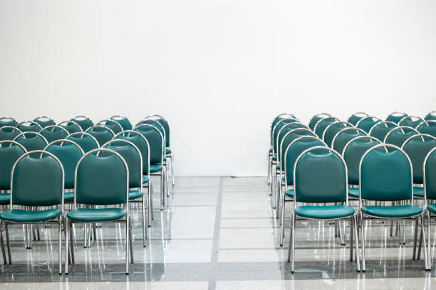 Many empty green leather chairs in a wide hall room, lights on. A floor reflect the objects. Give a feeling of emptiness.