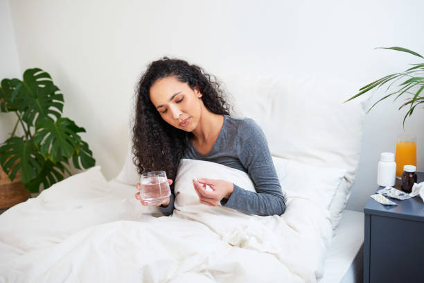 a young multi-ethnic woman takes a tablet in bed while sick with flu - multi vitamine stockfoto's en -beelden