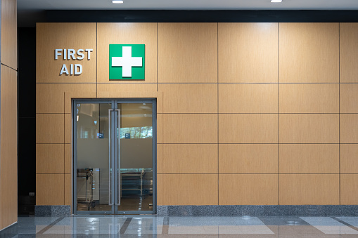 First Aid room inside a building. Decorated with pain light brown wall, push and pull door at an entrance and left open door to a scary walk way at a background with dim light.