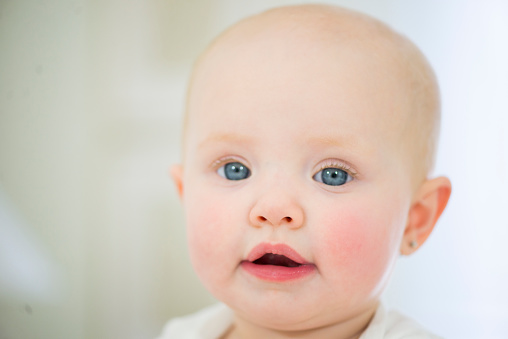 Closeup portrait of perfect, beautiful baby girl with pink lips and cheeks, blue eyes and peach fuzz hair. She is looking at the camera with a slight smile.