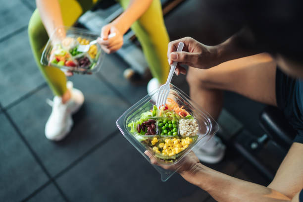 Top view Asian man and woman healthy eating salad after exercise at fitness gym. Top view Asian man and woman healthy eating salad after exercise at fitness gym. Two athlete eating salad for health together. Selective focus on salad bowl on hand. meal stock pictures, royalty-free photos & images
