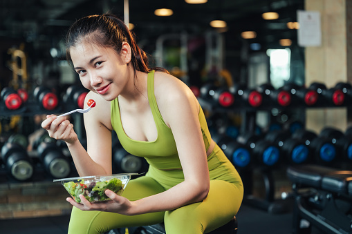 Beautiful asian woman in sportswear healthy eating salad after exercise at fitness gym. Healthy lifestyle. Young female smiling with holding salad bowl and looking at camera.