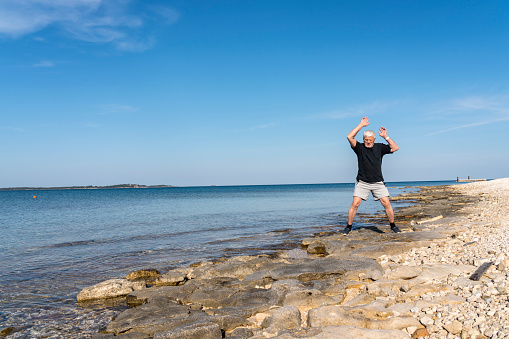 Side view of senior men exercising on flat rocks at the blue tranquil sea on sunny morning.