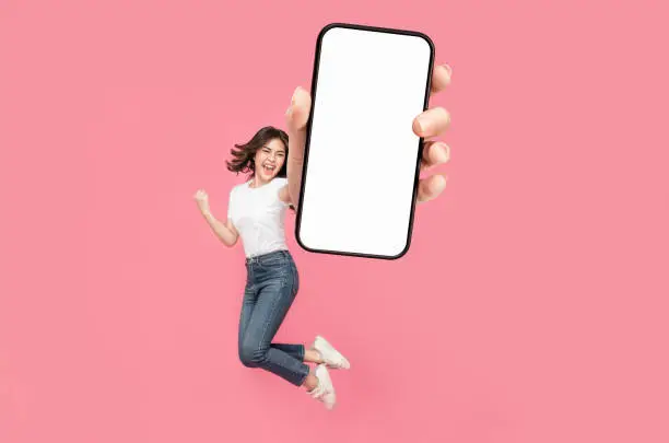 Photo of Full length of cheerful Asian woman jumping and smiling in air with showing cellphone blank screen with empty space for mobile app on screen.