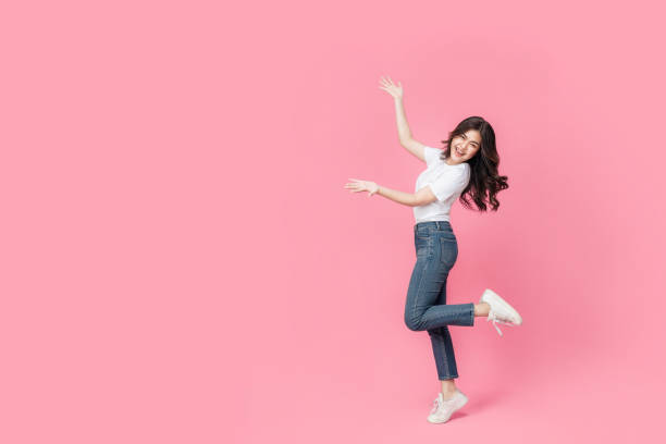 An asian woman in a casual outfit, hopping off with her arm presenting something in pink background. stock photo