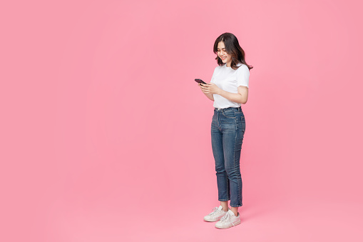 A full length of a cheerful asian woman in a white t-shirt with holding a mobile phone, enjoying the phone as she has got something exciting in isolated pink background.