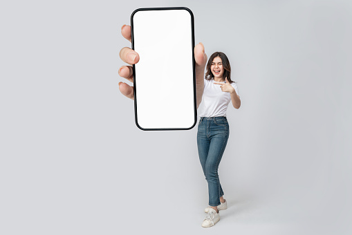 Full length of cheerful Asian woman smiling with showing cellphone blank screen with empty space for mobile app on screen. Isolated in studio white background. Creative collage.