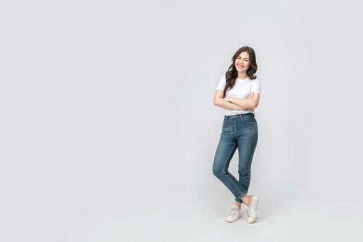Full length of confident asian woman smiling in a casual outfit in a white t-shirt and jeans, standing with her arm and leg crossed on isolated white background.