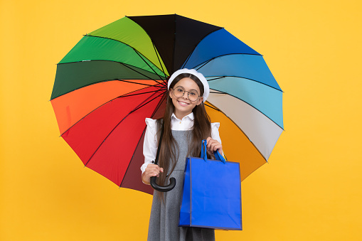 happy teen girl in glasses and beret under colorful umbrella for rain protection in autumn season hold shopping bag, autumn sale.