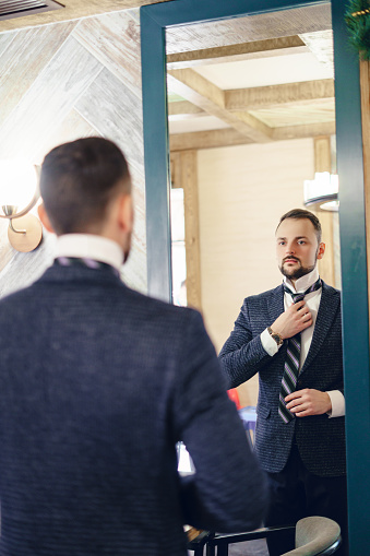 A young man with a small beard adjusts his tie while looking in a large mirror.