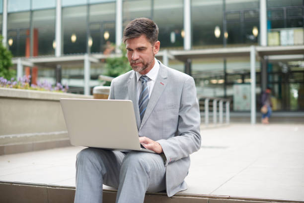 istock Business man typing on a laptop outdoors in a city booking a flight online using 5g internet connection. Corporate executive male sitting outside an office building on a break reading an email 1424010719