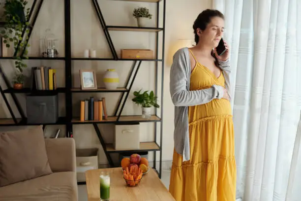 Serious pregnant young woman standing at window and talking on phone