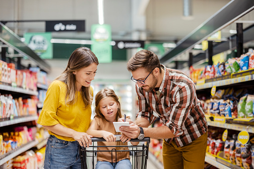 Family with shopping list buying food at grocery store or supermarket - sale, consumerism and people concept