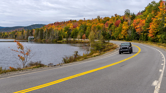 Rangeley, Maine, USA - September 29, 2021: A SUV driving along Beaver Pond on winding Route 17, part of Rangeley Lake Scenic Byway, on a cloudy but colorful Autumn morning.