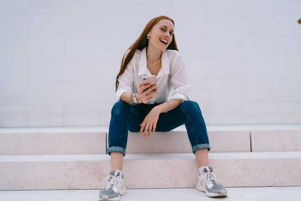 istock Cheerful young woman using smartphone while sitting on stairs 1424013365