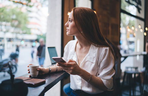 Pensive female in white blouse using cellphone while sitting at narrow table in coffee shop and looking through window in afternoon