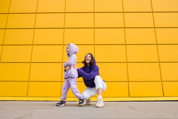 Woman in purple hoody have fun with cute girl 4-5 years old. Mommy and little daughter on yellow mall wall background. Childhood concept of love for family and fatherhood on Mother's Day stock photo