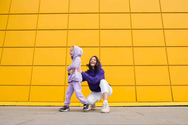 mulher de capuz roxo divirta-se com uma menina bonita de 4 a 5 anos. mamãe e filha no fundo da parede amarela do shopping. conceito infantil de amor à família e paternidade no dia das mães - child 4 5 years laughing little girls - fotografias e filmes do acervo