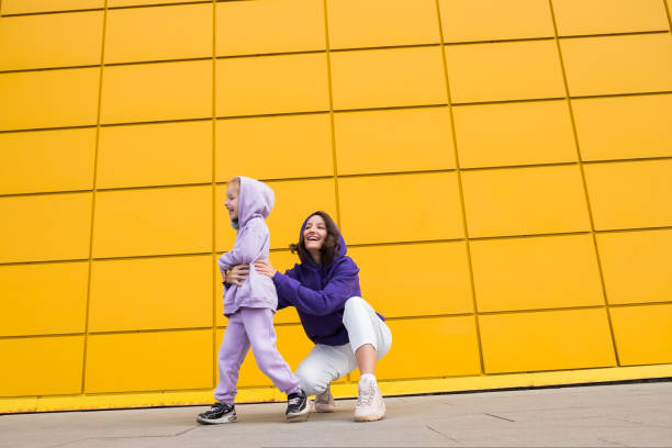 mulher de capuz roxo divirta-se com uma menina bonita de 4 a 5 anos. mamãe e filha no fundo da parede amarela do shopping. conceito infantil de amor à família e paternidade no dia das mães - child 4 5 years laughing little girls - fotografias e filmes do acervo