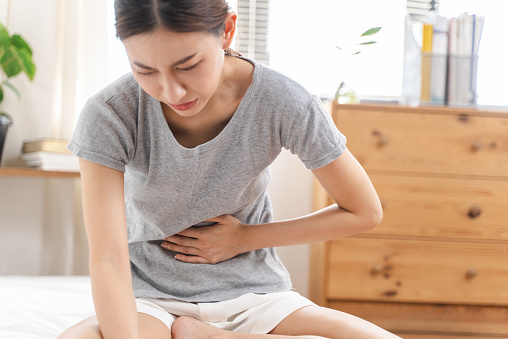 Close-up beautiful young Asian woman suffering from stomachache or menstruation pain. Unhealthy teenage girl sitting in bedroom, touching her belly feeling pain and unwell.
