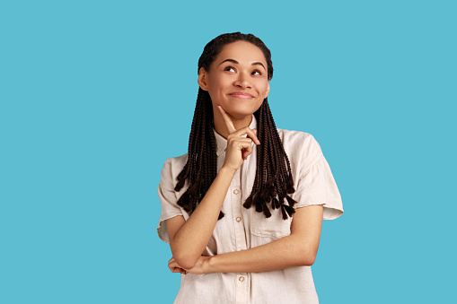 Portrait of pretty woman with dreadlocks, having satisfied expression, keeps finger on chin, concentrated away, thinks about pleasant moment in life. Indoor studio shot isolated on blue background.