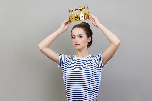 Portrait of serious confident woman wearing striped T-shirt and golden crown looking with arrogance and confidence, privileged status. Indoor studio shot isolated on gray background.