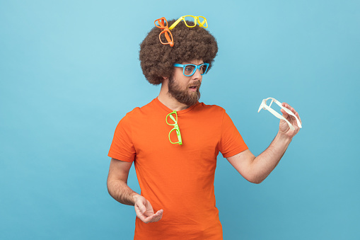 Portrait of funny man with Afro hairstyle wearing orange T-shirt choosing colorful eyeglasses, holding white spectacles, choosing the best color. Indoor studio shot isolated on blue background.