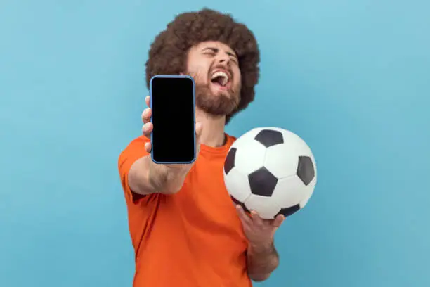 Photo of Man standing with soccer ball and showing mobile phone with empty display, betting and winning.