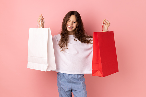 Portrait of happy satisfied little girl wearing white T-shirt holding shopping paper bags, excited with mall discounts, good purchases. Indoor studio shot isolated on pink background.