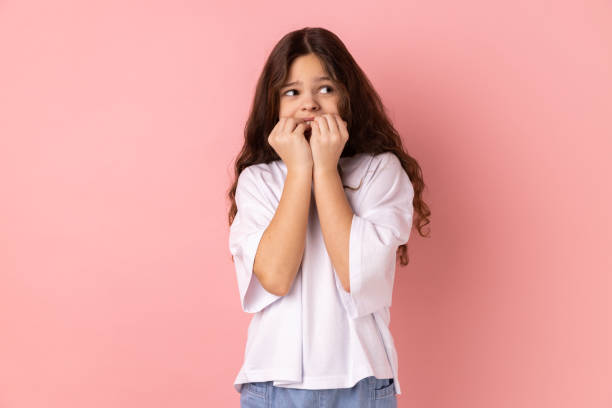 Little girl biting nails, terrified about problems, suffering phobia, anxiety disorder. Troubles and worries. Portrait of little girl wearing white T-shirt biting nails, terrified about problems, suffering phobia, anxiety disorder. Indoor studio shot isolated on pink background. nail biting stock pictures, royalty-free photos & images