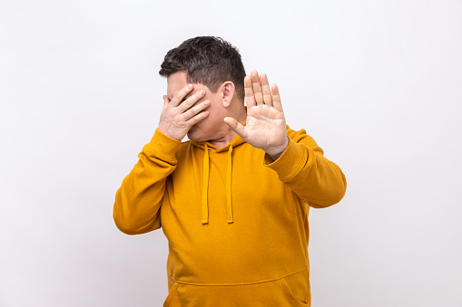 Portrait of confused man covering eyes and raising hand to stop, feeling stressed afraid, refusing to watch scary content, wearing urban style hoodie. Indoor studio shot isolated on white background.