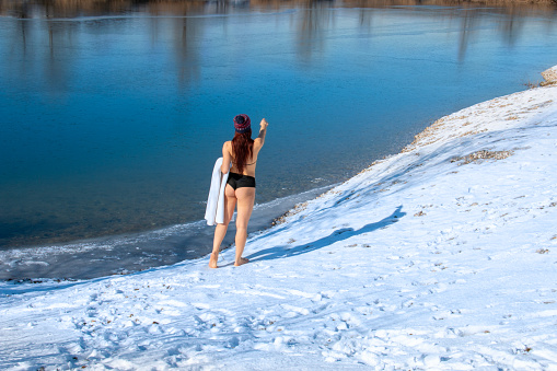 Back view beautiful woman in swimwear walking barefoot on snow in winter towards the icy lake landscape