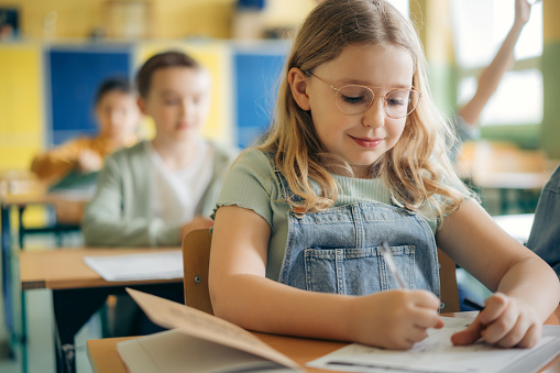 Portrait of a cute elementary schoolgirl sitting at desk in classroom. Small girl sitting at classroom desk and looking at camera.