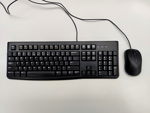 Unbranded black QWERTY keyboard with unbranded black mouse and cables on a white office desk. There are no people in the shot.